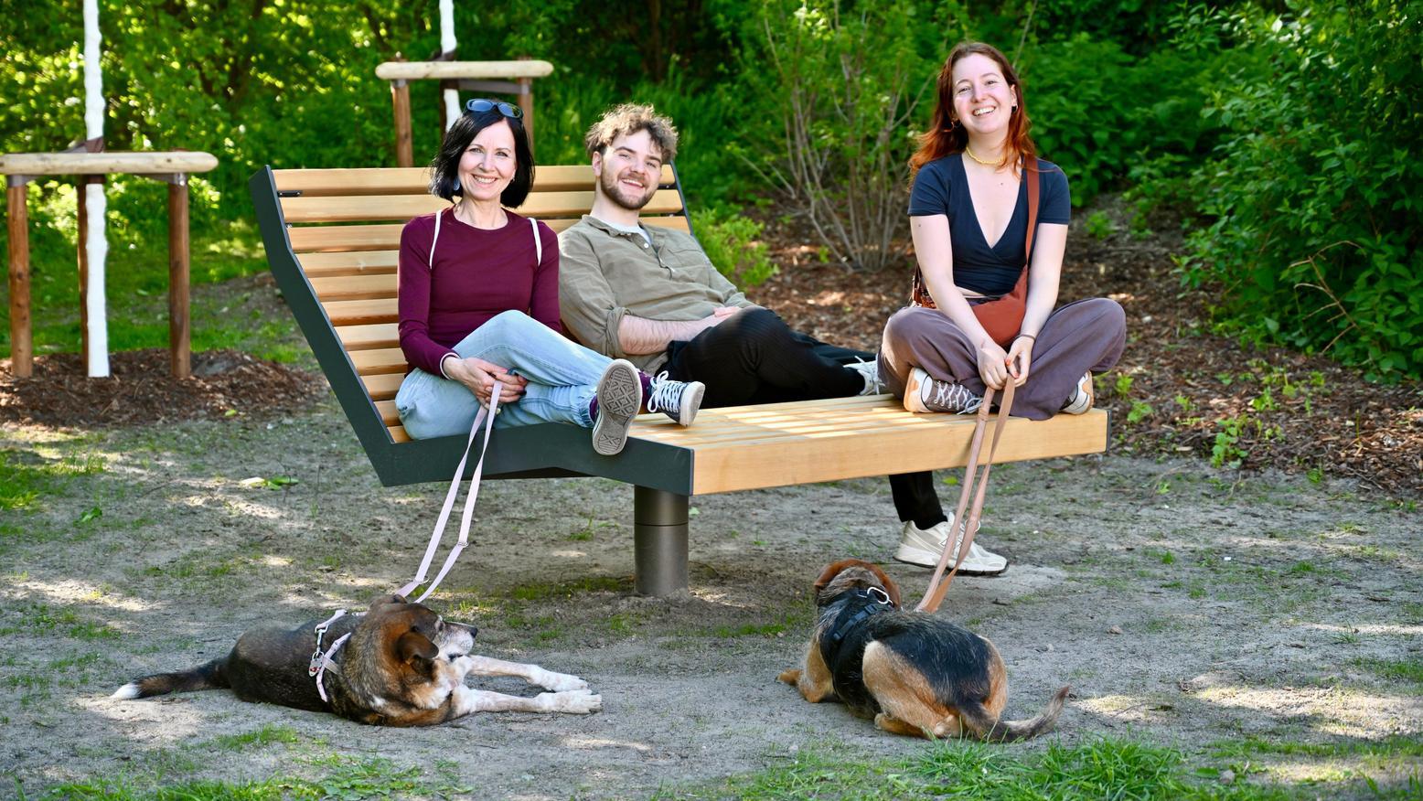 Drei Personen sitzen auf einer Bank und zwei Hunde liegen vor ihnen auf dem Boden.