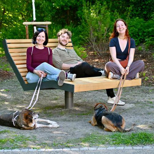 Drei Personen sitzen auf einer Bank und zwei Hunde liegen vor ihnen auf dem Boden.
