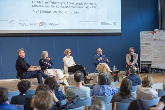 Fünf Personen sitzen auf Stühlen vor einer Leinwand und diskutieren.
