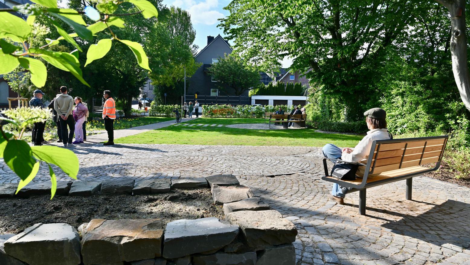 Ein Mann sitzt auf einer Bank, im Hintergrund ist eine Parkfläche und Menschen stehen auf einer Wiese. 
