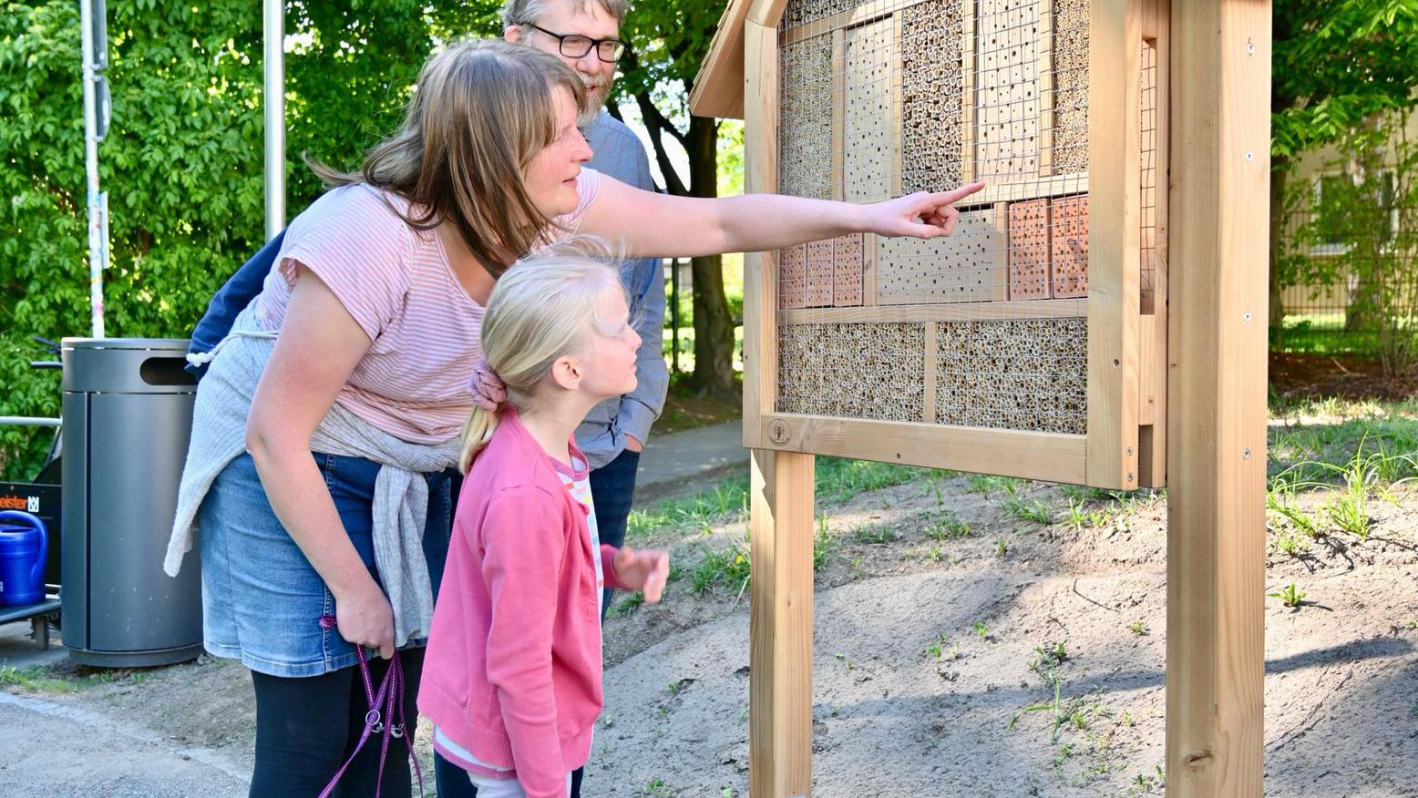 Eine Familie mit Mädchen und Hund schauen sich ein Insektenhotel an. 
