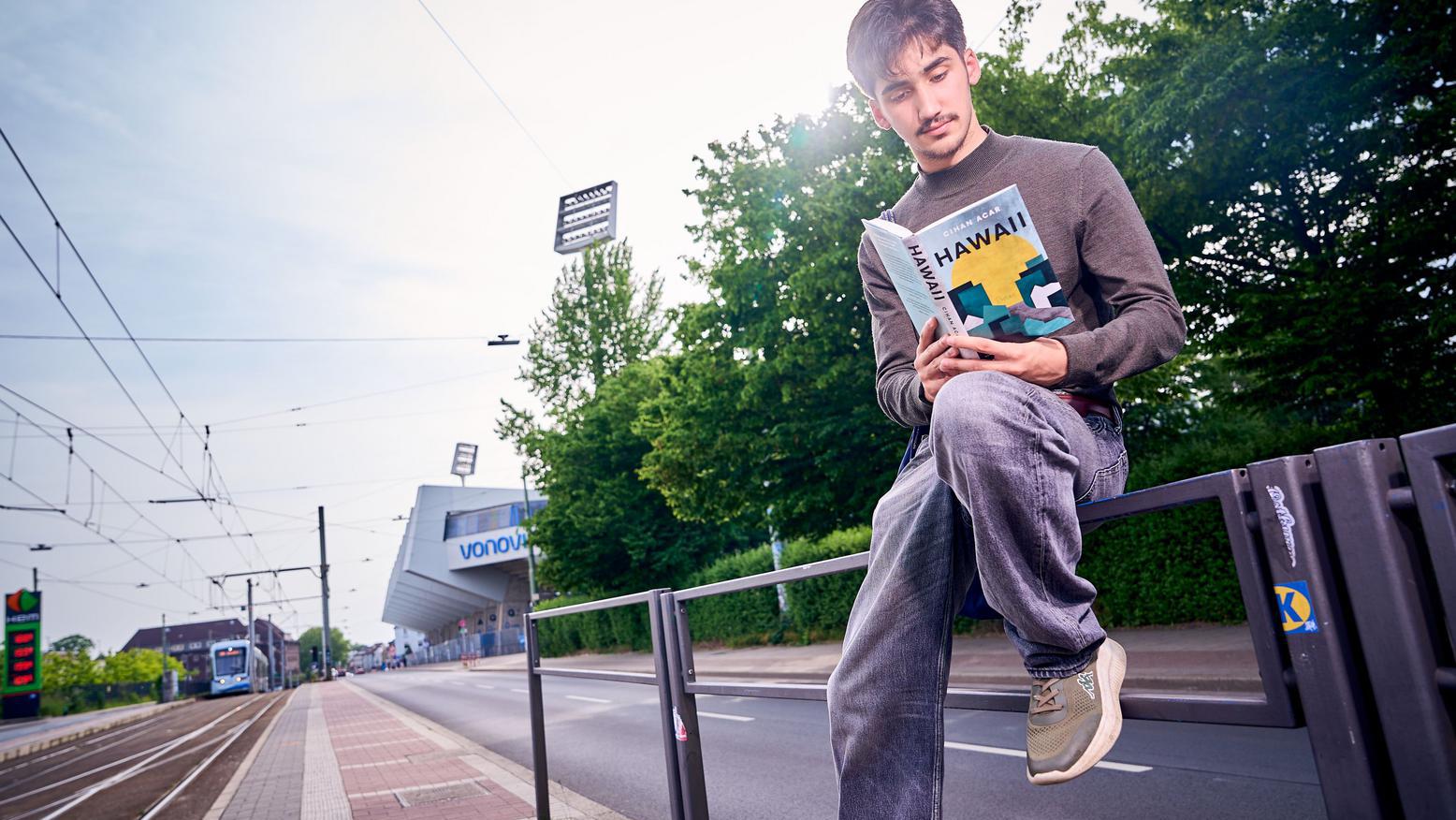 Ein Mann sitzt auf dem Geländer an einem Bahnsteig und liest das Buch "Hawaii"
