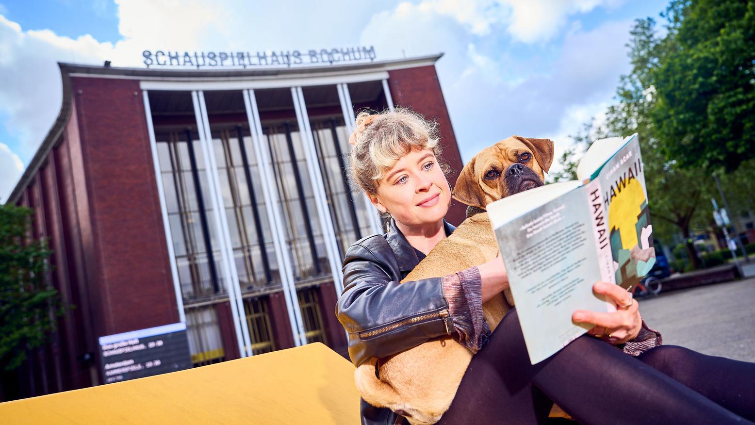 Eine Frau sitzt mit ihrem Hund im Arm vor den Schauspielhaus Bochum und liest das Buch "Hawaii"