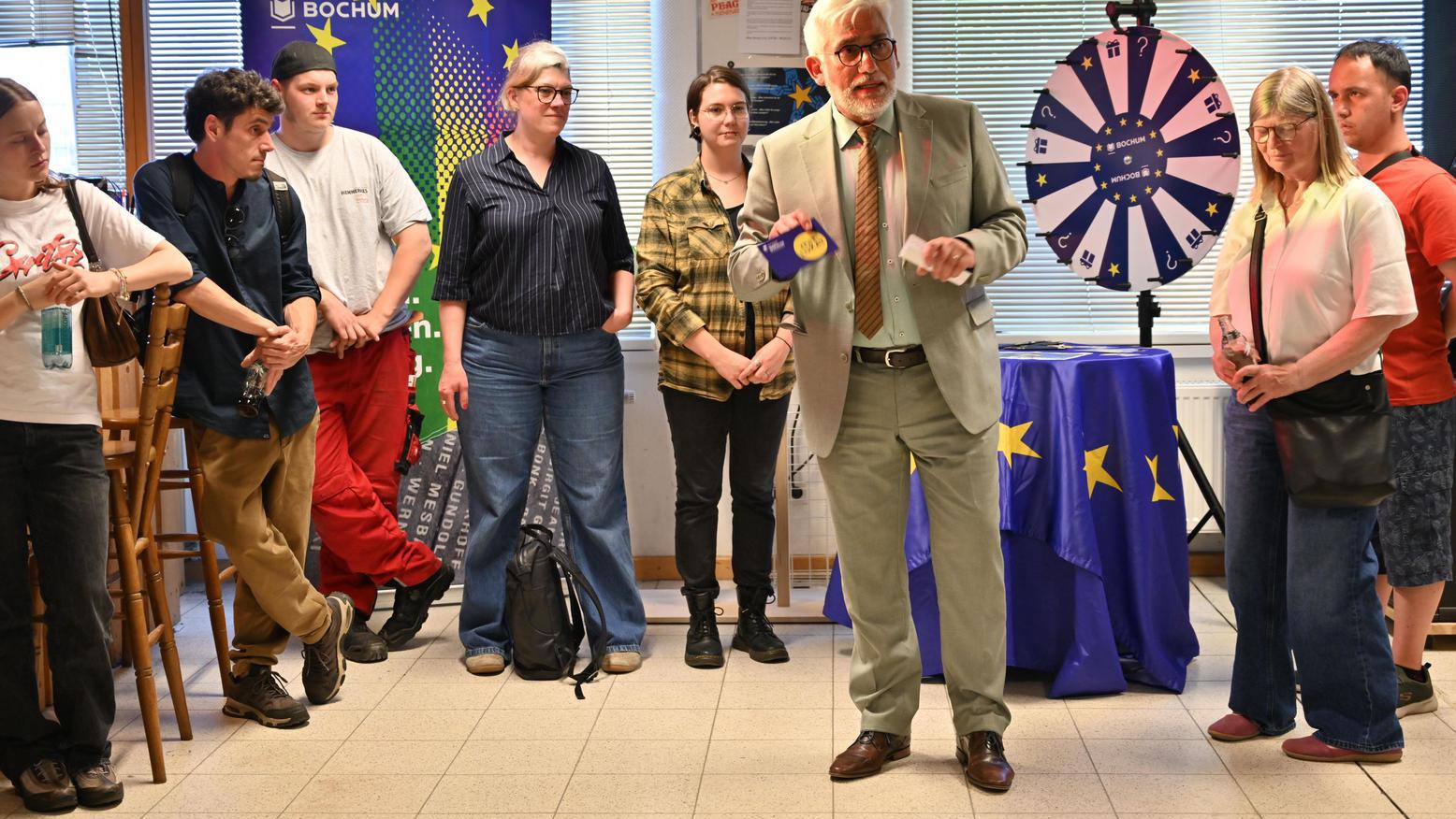 Oberbürgermeister Jörg Lukat spricht bei einer Veranstaltung vor einer Gruppe von Besucherinnen und Besuchern. Im Hintergrund stehen ein Europa-Banner und ein Glücksrad.