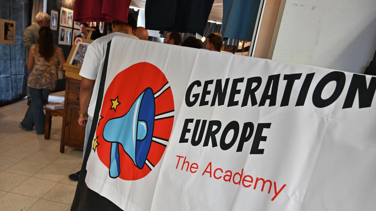 Banner von "Generation Europe – The Academy" in einem Ausstellungsraum; im Hintergrund sind Besucherinnen und Besucher sowie eine Fotoausstellung zu sehen.