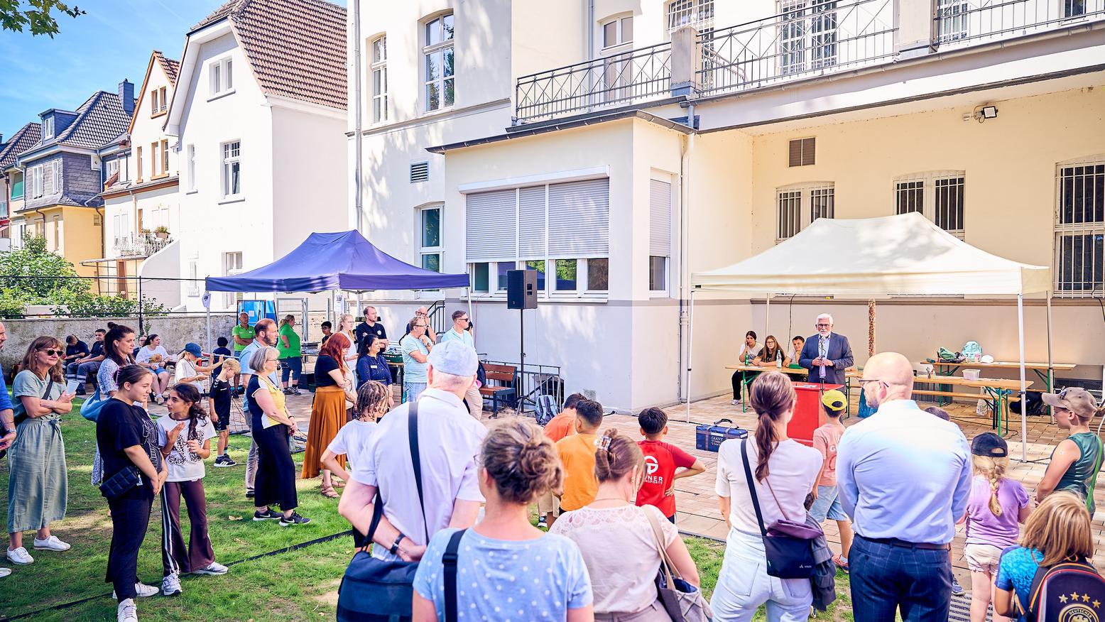 Besucherinnen und Besucher verfolgen eine Ansprache bei der Eröffnung eines Außengeländes vor einem Gebäude.