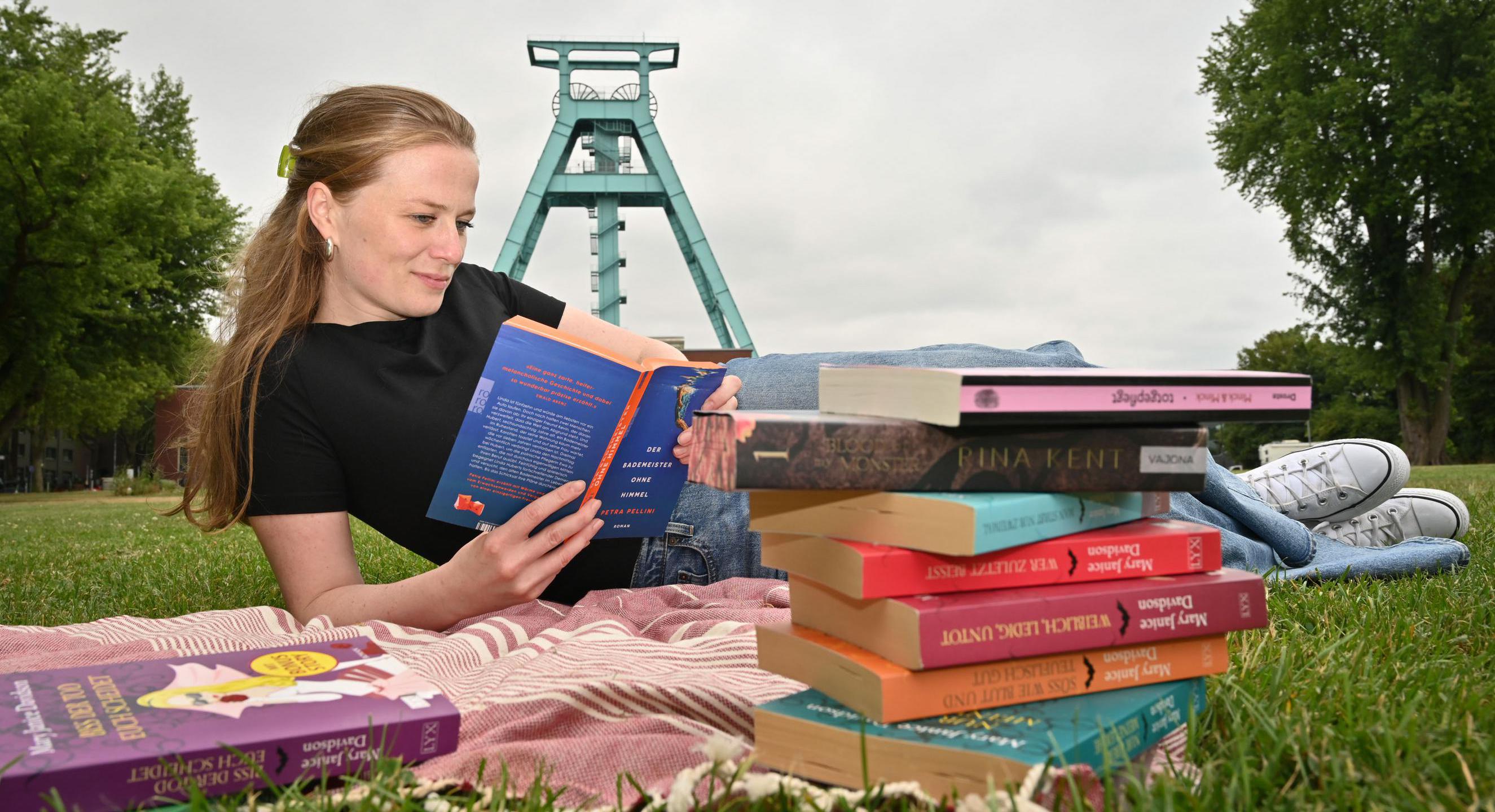 Person liest auf einer Decke im Freien ein Buch. Im Vordergrund liegen mehrere Bücher, im Hintergrund ist ein Fördergerüst zu sehen.