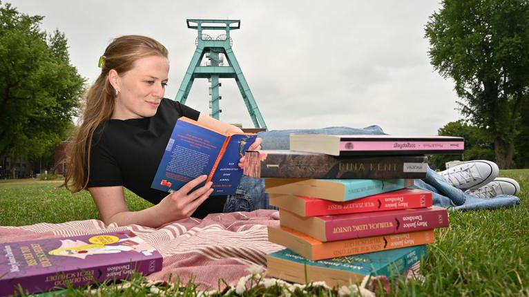 Person liest auf einer Decke im Freien ein Buch. Im Vordergrund liegen mehrere Bücher, im Hintergrund ist ein Fördergerüst zu sehen.