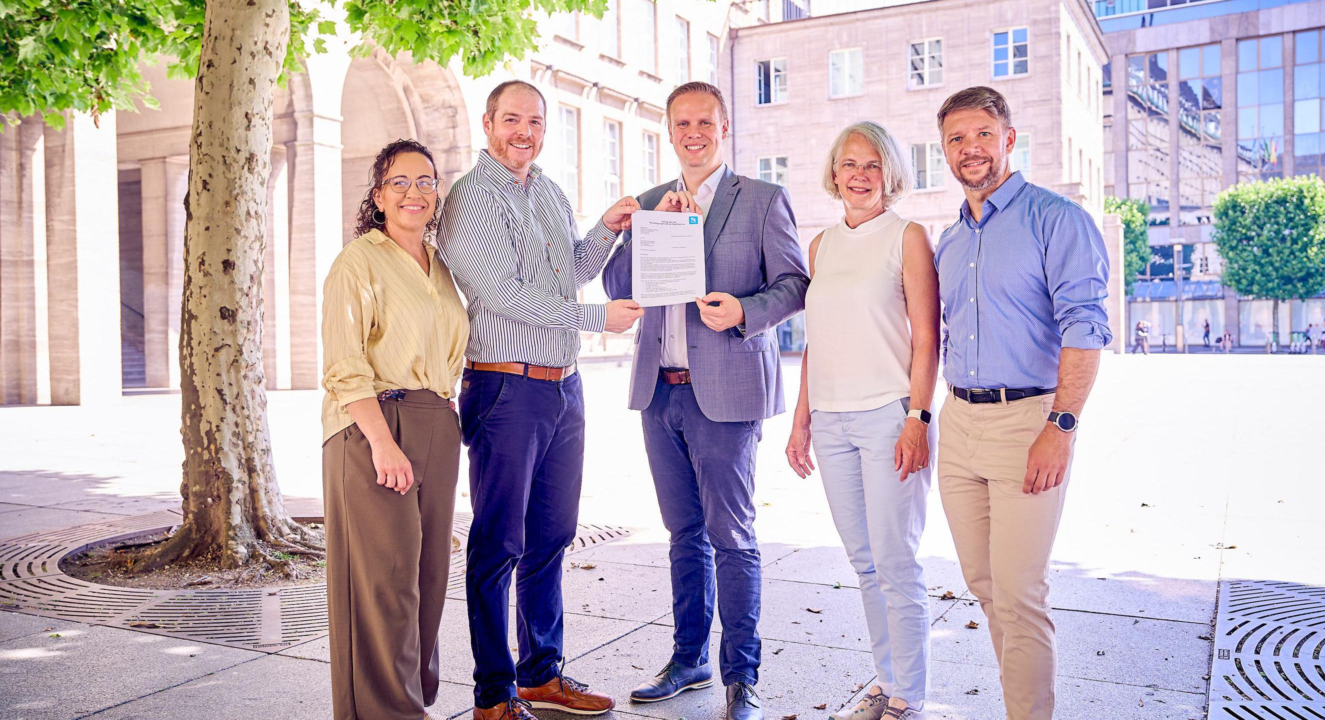 Fünf Personen vor dem städtischen Rathaus Bochum, die beiden Personen in der Mitte des Bildes halten den Vertrag zum TK-Verfügungsfonds in den Händen.