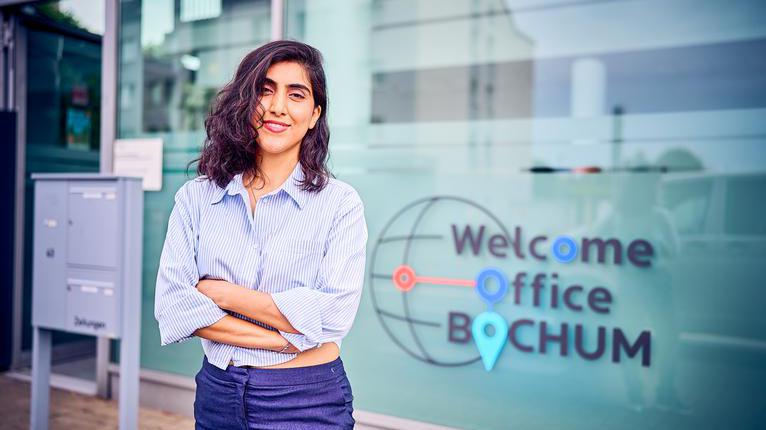 Eine junge braunhaarige Frau in Jeans und Bluse vor der Außenfassade des Welcome Office Bochum.