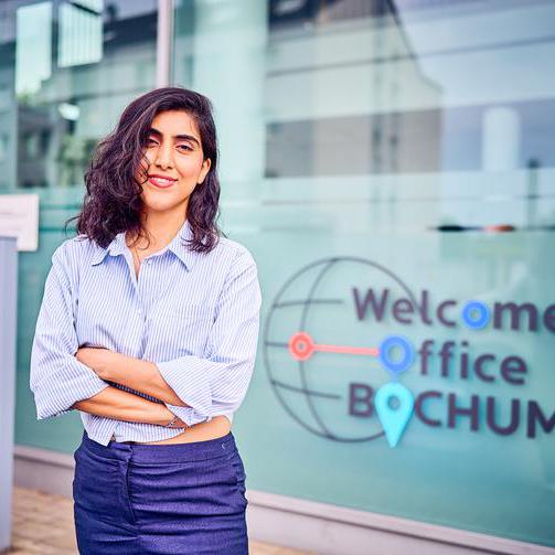 Eine junge braunhaarige Frau in Jeans und Bluse vor der Außenfassade des Welcome Office Bochum.