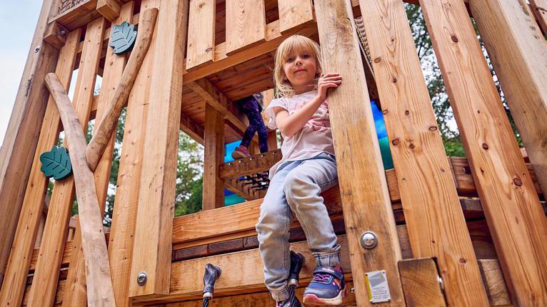 Ein Kind sitzt in einem Turm aus Holz. Es sitzt an einem Durchgang und lächelt leicht in die Kamera. Im Hintergrund klettert ein weiteres Kind durch den Turm.