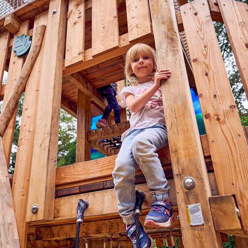 Ein Kind sitzt in einem Turm aus Holz. Es sitzt an einem Durchgang und lächelt leicht in die Kamera. Im Hintergrund klettert ein weiteres Kind durch den Turm.