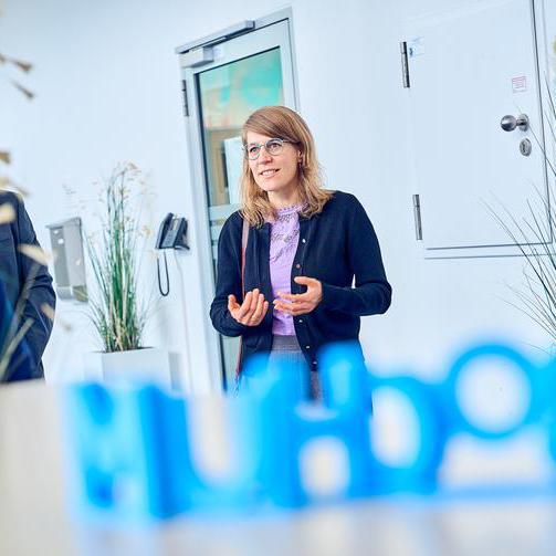NRW Ministerin Schäffer unterhält sich. Sie hält ihr Hände vor ihrem Oberkörper. Sie lächelt. Im Vordergrund ein blauer Bochum Schriftzug, spiegelverkehrt. Im Hintergrund ein weißer. moderner Raum.